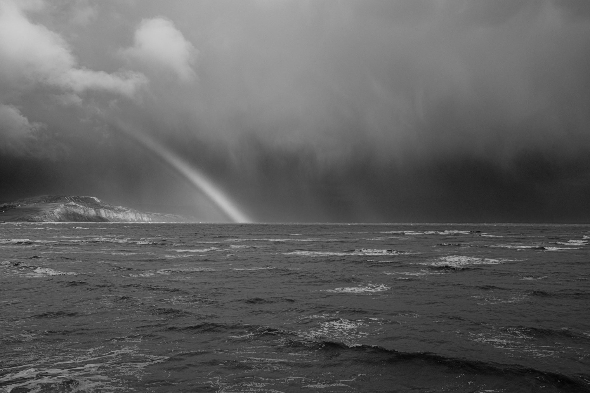 Rainbow in sunlight and a stormy sky over the coast from Church Cliff Walk Lyme Regis ACROS R 27_03_24 1 mono